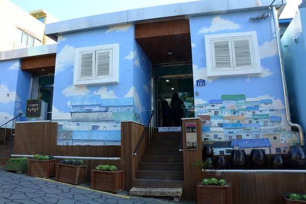 Bước đến đầu làng, bạn sẽ ấn tượng ngay với ngôi nhà màu xanh sơn vẽ các công trình đặc trưng ở Gamcheon. Đây cũng là nơi du khách bắt đầu hành trình khám phá làng văn hóa theo tấm bản đồ Gamcheon lấy tại quầy thông tin.