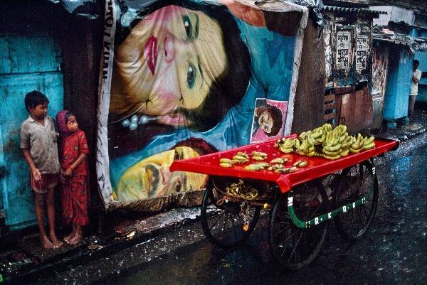 Hai đứa trẻ nép vào nhau trú mưa trong một mái hiên, bên ngoài là xe chuối chỉ có vài ba nải ở Bombay, Ấn Độ