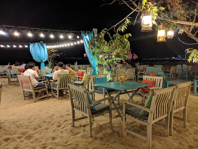 19h30: Nhâm nhi đồ uống ở Surf Bar Nghỉ ngơi sau một ngày khám phá thành phố biển, Surf Bar là điểm đến lý tưởng tiếp theo với điểm cộng là những bộ bàn ghế gỗ đặt ngay trên nền cát, nằm trên đường ven bờ biển Xuân Diệu. Ngồi thưởng thức những món đồ uống tại quán, du khách có thể tận hưởng không gian thoáng đãng với những làn gió biển mát rượi, tiếng sóng vỗ rì rào. Một số đồ uống như cocktail, nước ép đều có giá dưới 40.000 đồng. Ảnh: Vy An.